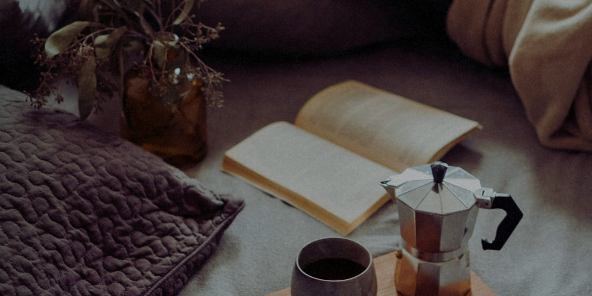 A softly lit bedside scene with an open notebook, a cup of tea, a textured throw blanket, and a small vase with dried stems, creating a calm, cozy atmosphere.