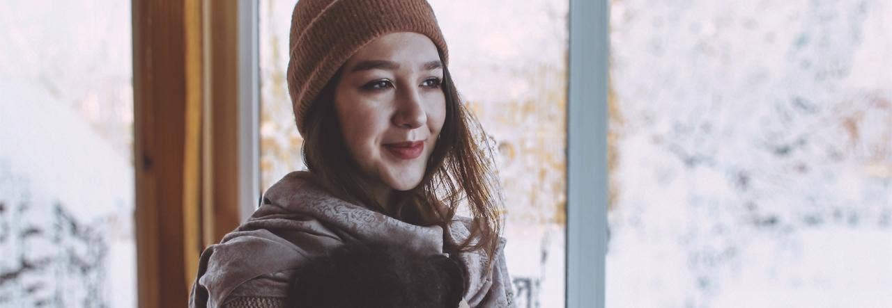 Woman wearing scarf, mittens and hat near a window, snow outside