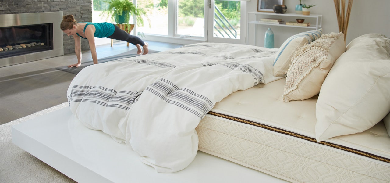 Bare mattress with bedding and pillows, woman exercising in the background