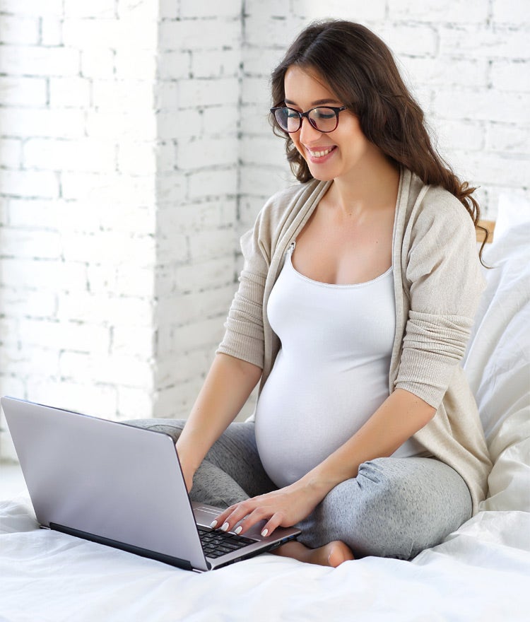 Pregnant woman sitting cross-legged on a bed, researching on a laptop 