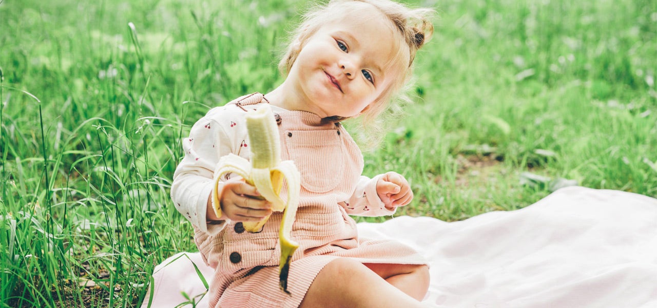 Toddler girl eating a banana on a blanket in the grass