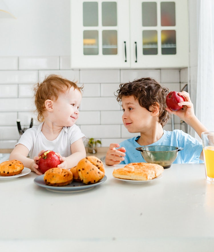 Two small children at the breakfast table, eating apples and scones