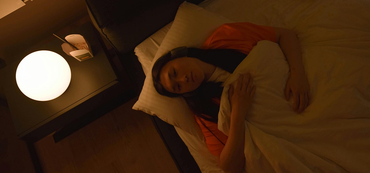person laying in bed with bedside lamp on and chinese food container on bedside table